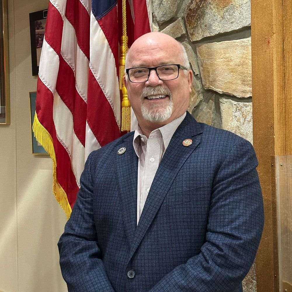Smiling man in a blue blazer standing beside an American flag indoors