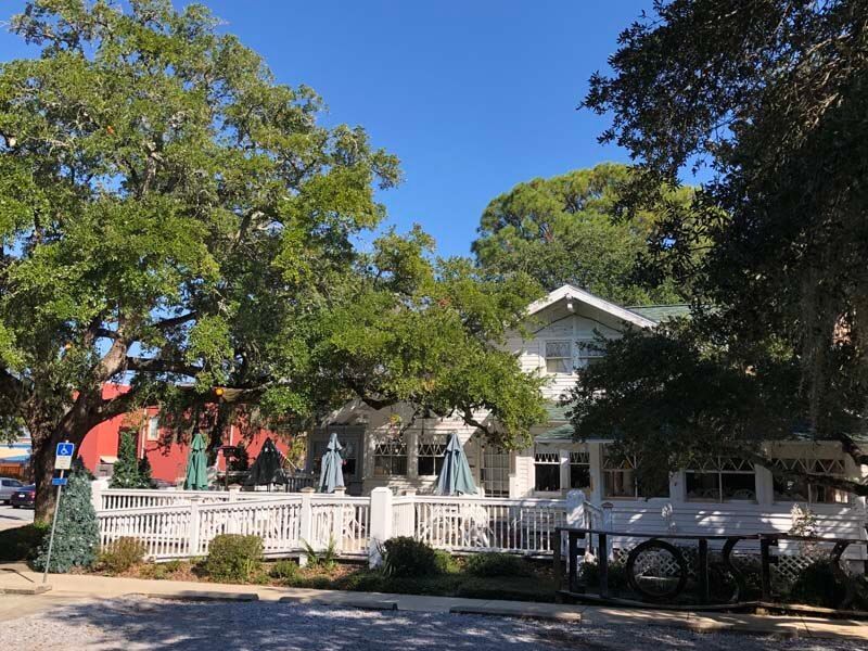 White house with porch and patio umbrellas behind trees and parked car on a sunny street