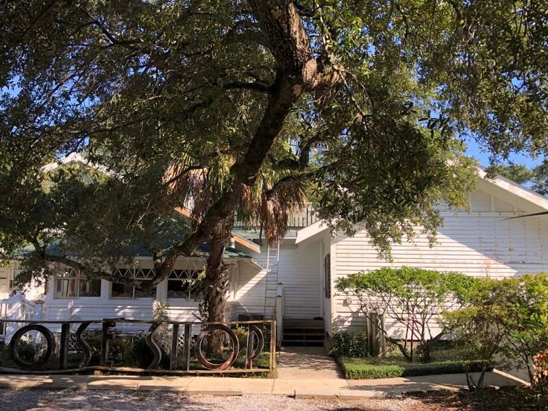 Tree shading a white house with a bicycle on the porch and a dirt path in front