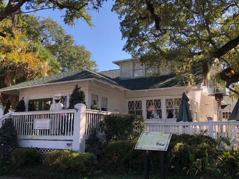 White house with porch and garden, surrounded by trees and white fencing in bright daylight