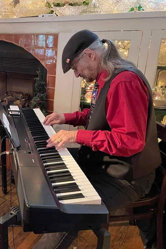 Man in a red shirt and black vest plays a piano indoors near a fireplace.