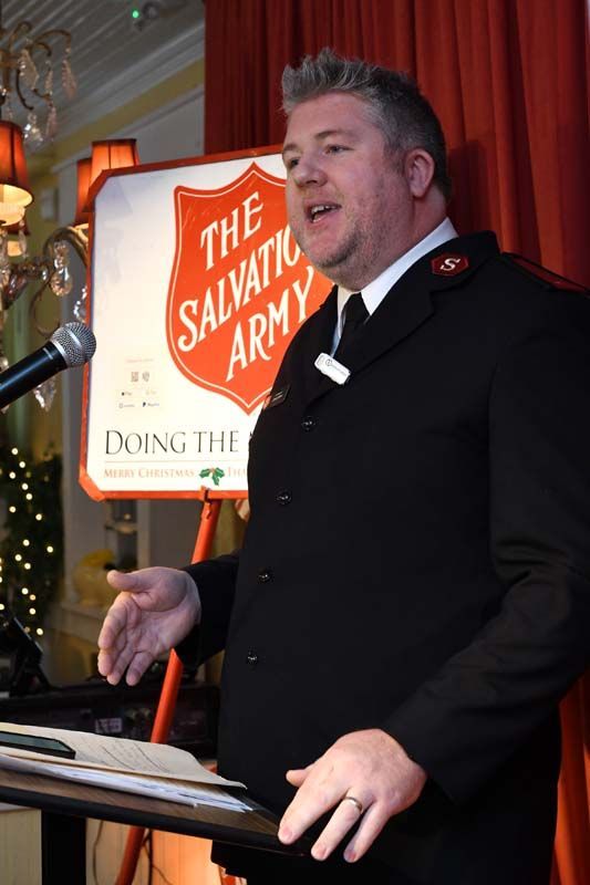 Man speaking at a podium beside a Salvation Army sign and microphone on a stage with red curtains