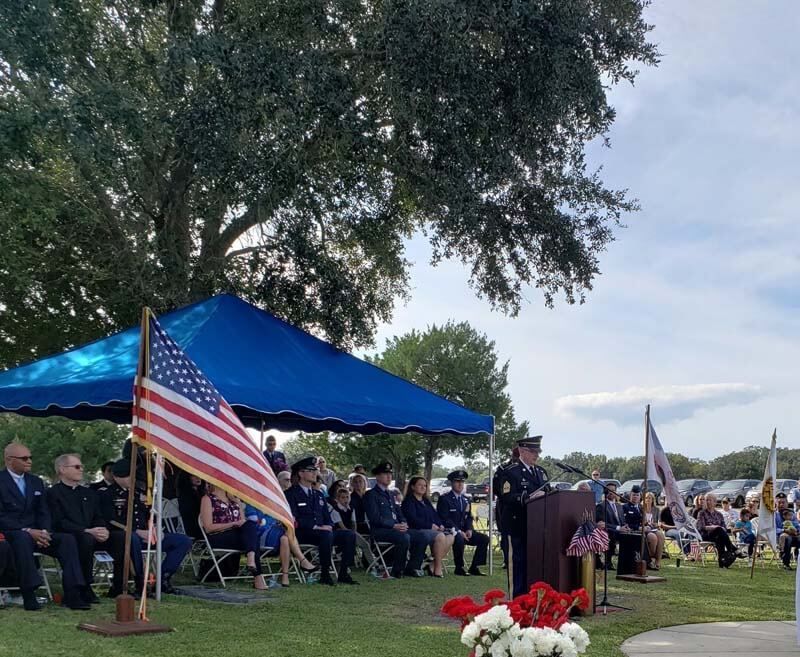 Outdoor memorial ceremony with speakers on a stage, American flags, and seated attendees under a blue canopy.