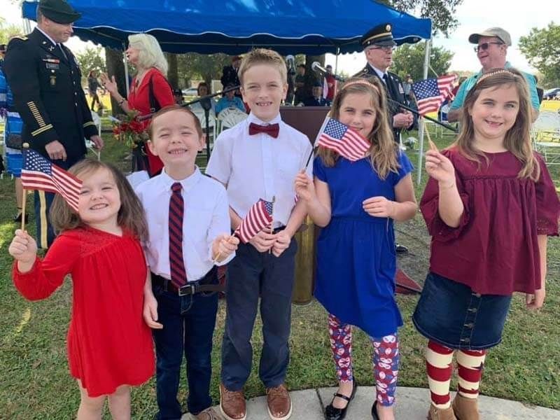 Six children posing with small American flags at an outdoor event, in red, white, and blue outfits.
