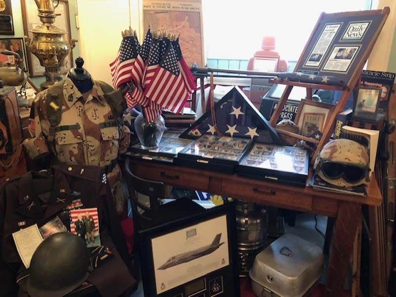 Patriotic military memorabilia display with flags, helmet, framed photos, and assorted artifacts on a desk and shelves