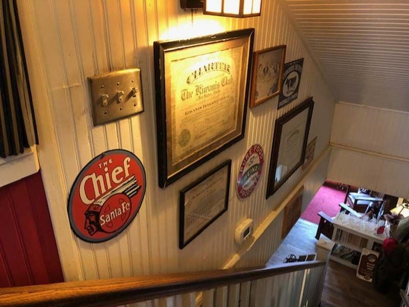 Stairwell wall with framed vintage signs and certificates, lit by a warm lamp, overlooking a bar below