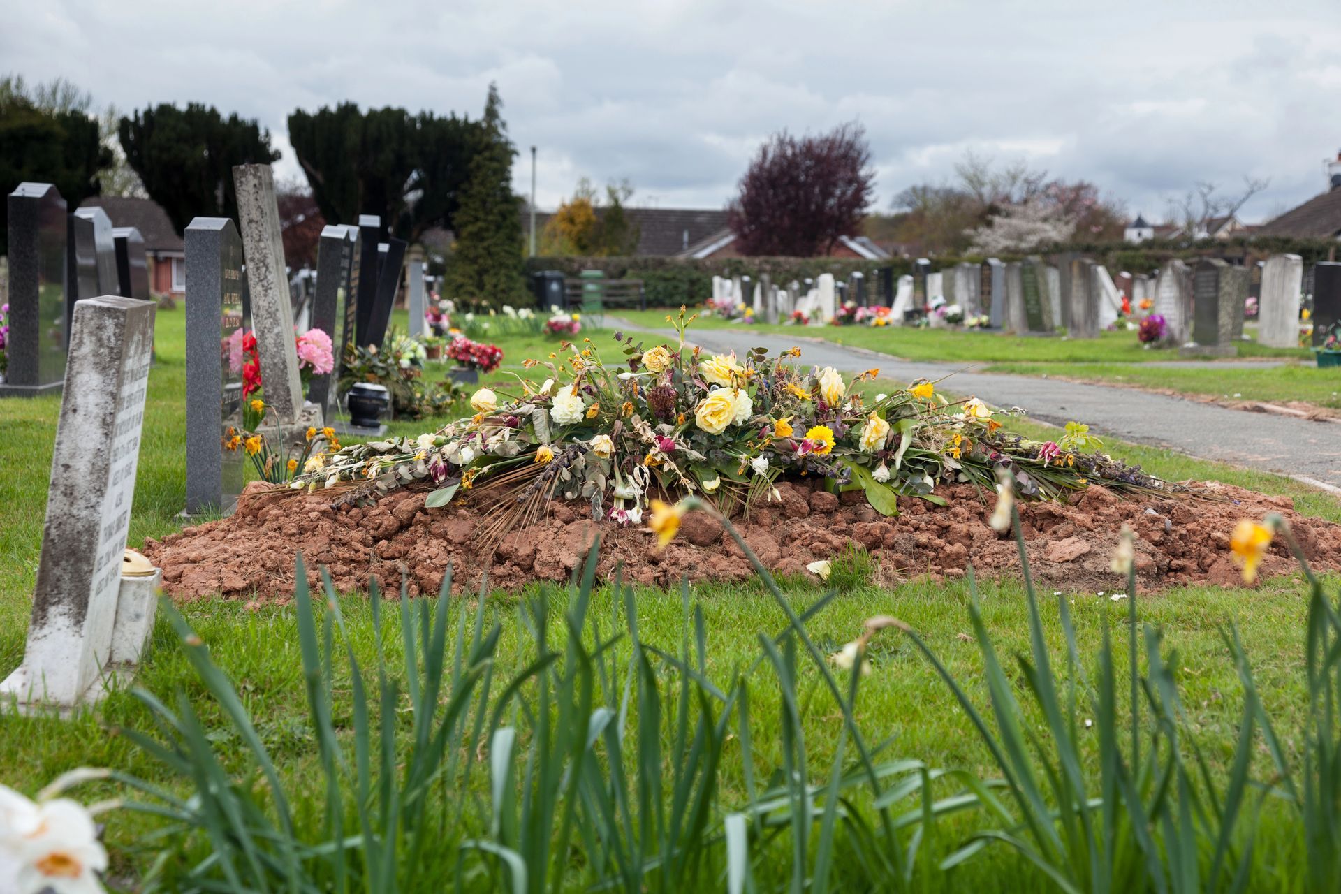 Tomba con terra fresca, fiori e lapidi in un cimitero. Cielo coperto.
