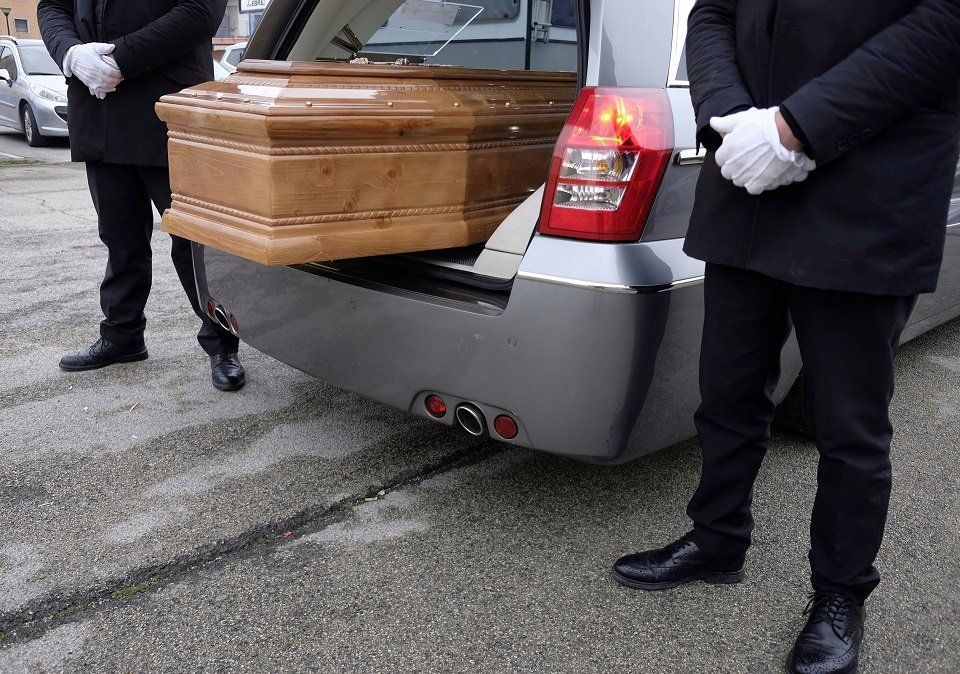 Carro funebre con bara; due persone in abito nero e guanti bianchi stanno lì vicino.