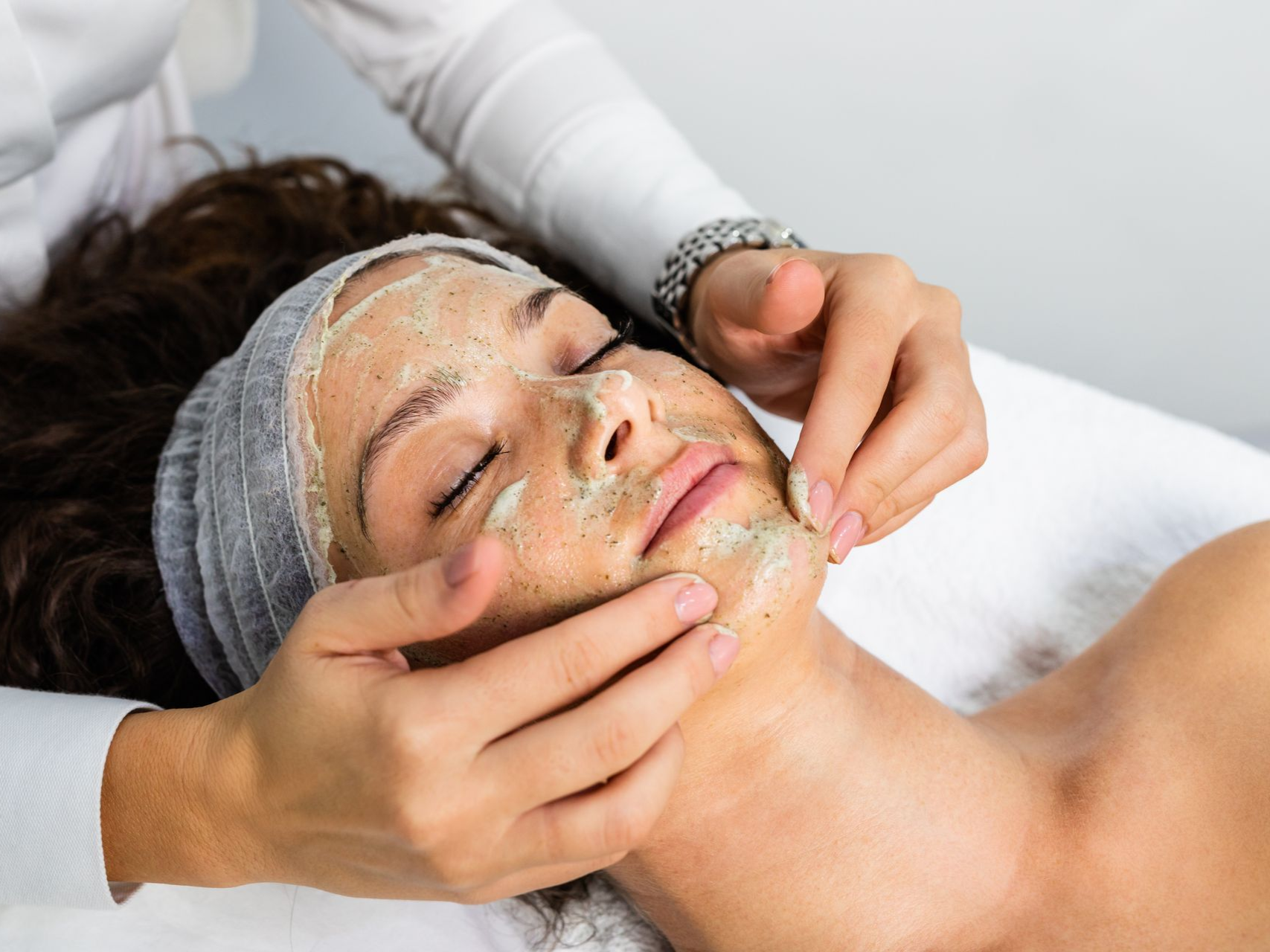 Facial treatment with masked face, hands gently massaging cheeks on a white spa bed