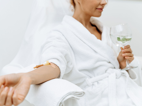 Woman in a white robe reclining on a spa lounge, holding a glass and extending her arm with a drip infusion