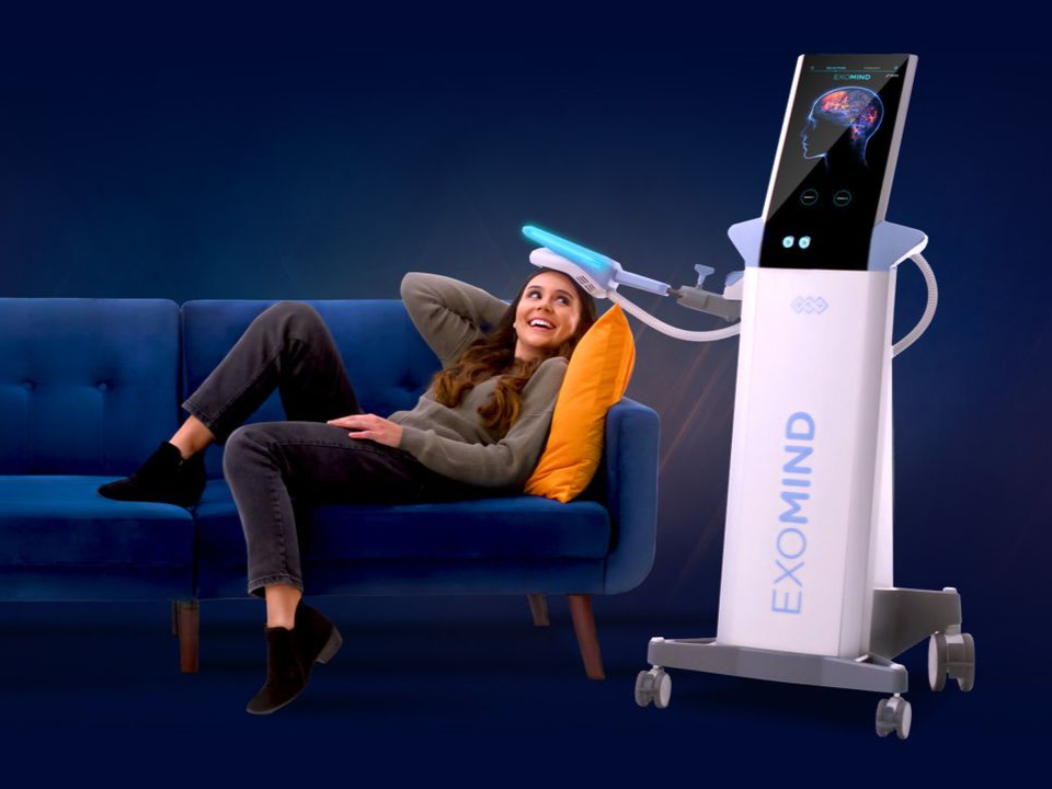 Person relaxing on a blue couch beside a white EXOMIND machine with a glowing screen.