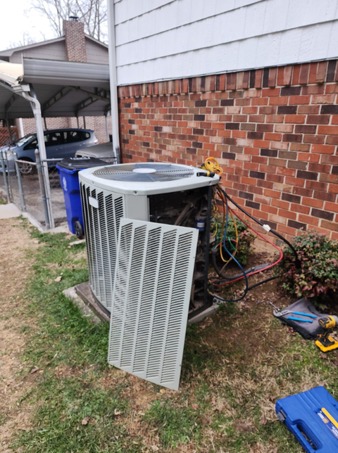 Outdoor AC unit beside a brick house with its front grille removed, exposing wires and components.