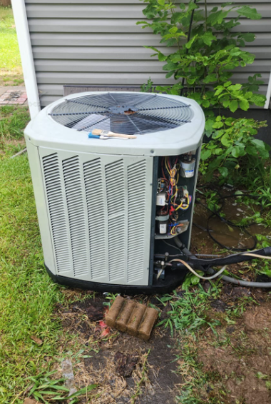 Outdoor air conditioner unit with side panel open, showing internal components beside a house wall and grass.