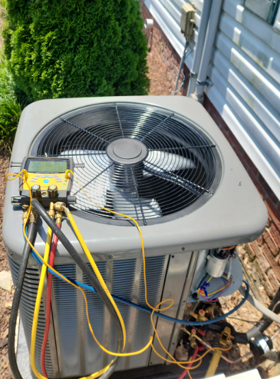 Outdoor HVAC unit with gauges and yellow hoses attached, beside a house wall and greenery