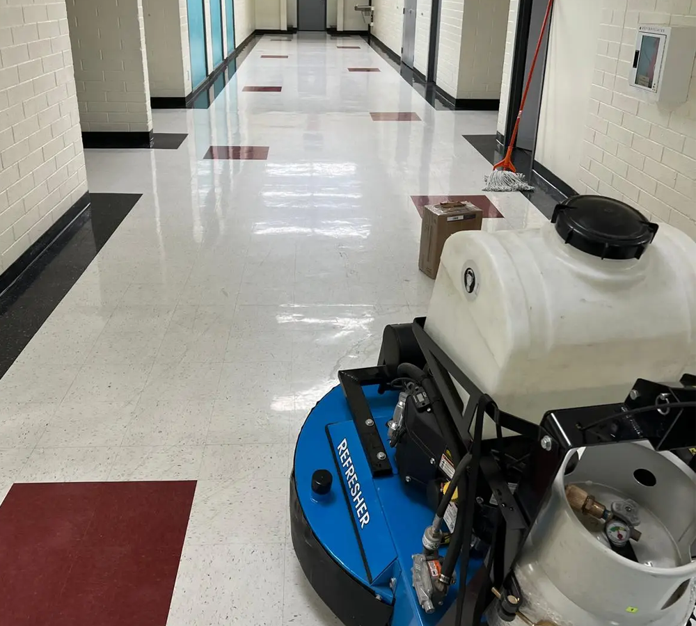 Blue floor-cleaning machine in a hallway with a white tile floor and red mats.