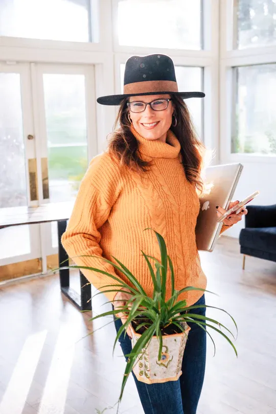 Smiling Tara Parsons wearing a fedora, holding a laptop and potted plant