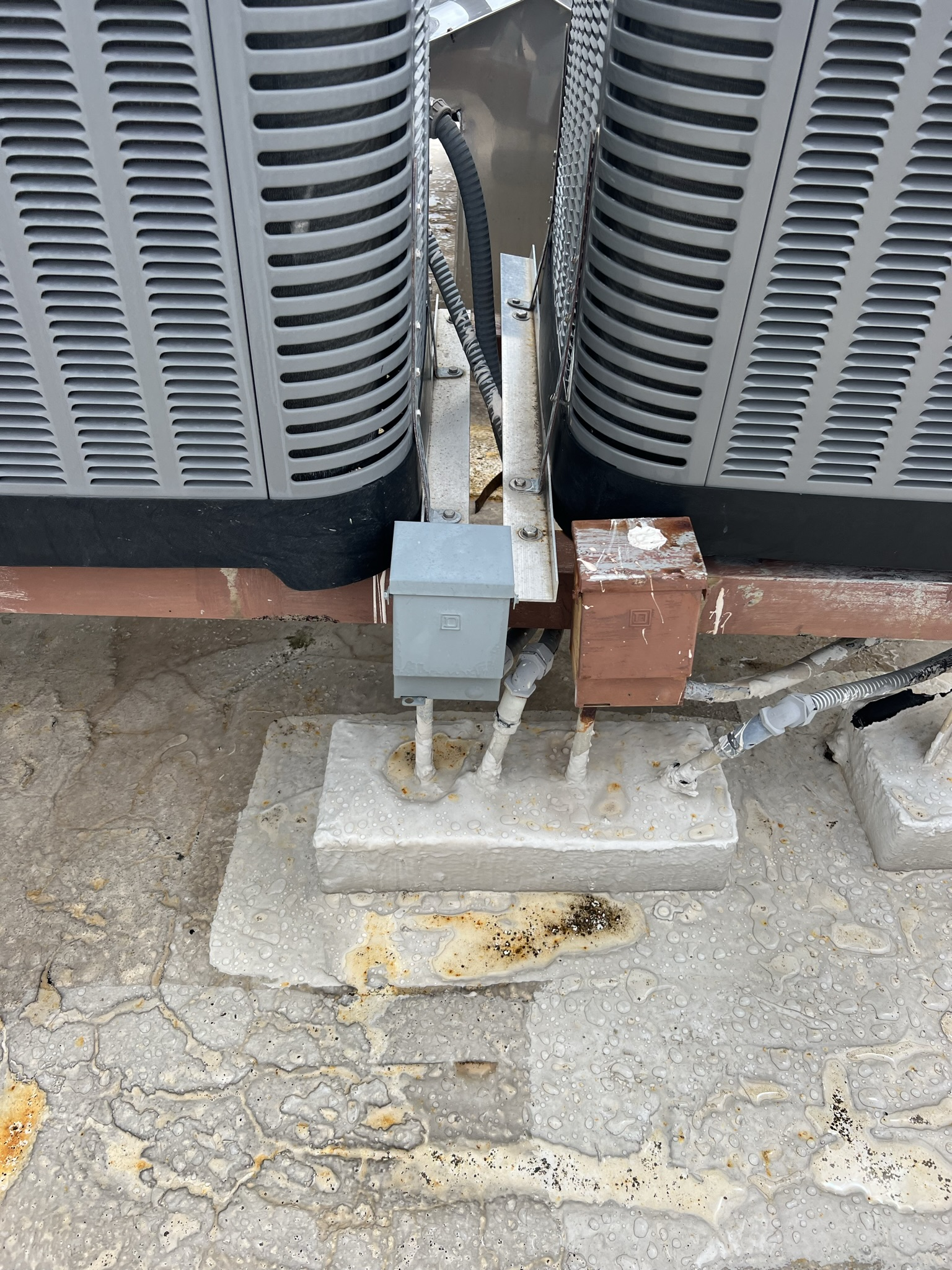 Two HVAC units with metal frames and a concrete base on a textured surface. Electrical boxes are visible.