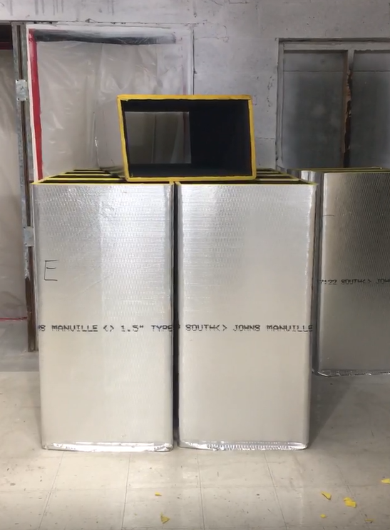 Silver insulated boxes with a yellow-framed vent against a plain wall, likely in a construction setting.
