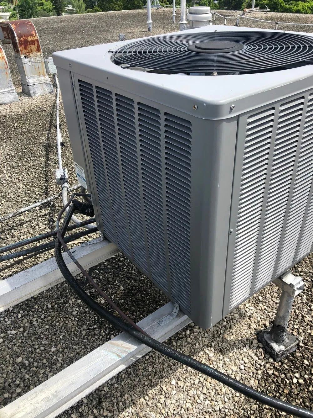Outdoor HVAC condenser unit with black fan on gravel beside metal rails and hoses
