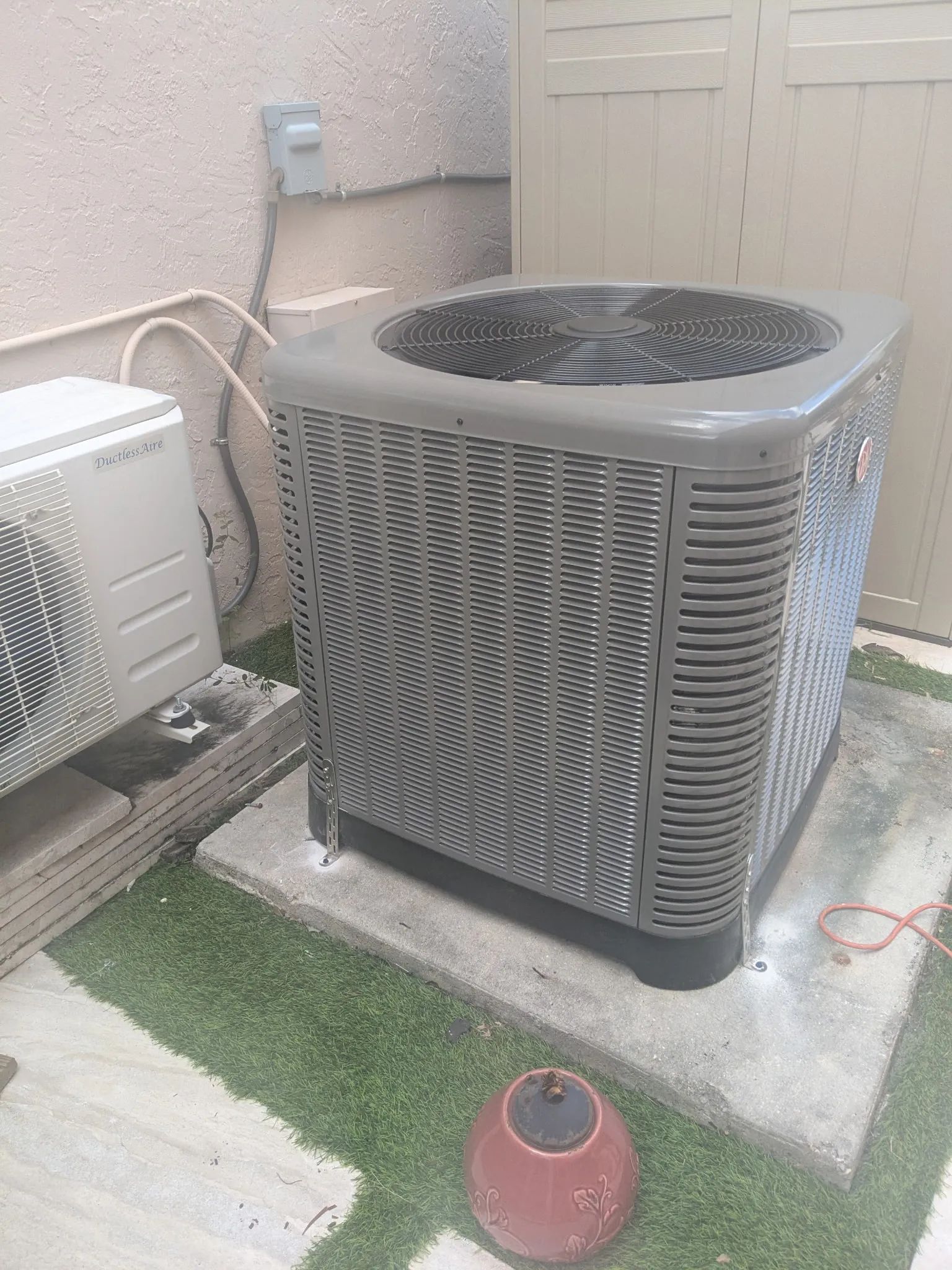 Outdoor air conditioner unit beside a beige wall and fence on a concrete pad, with a small red pot nearby.