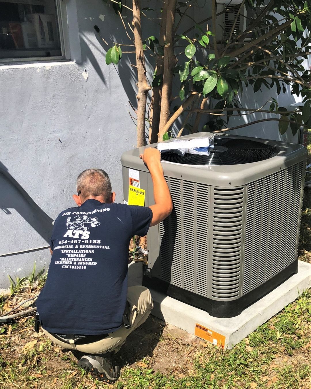 Worker crouches by an outdoor HVAC unit beside a building, adjusting a young tree