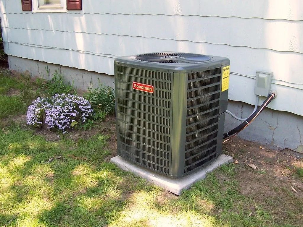 Outdoor air conditioner unit beside a house, near a small purple flower bush