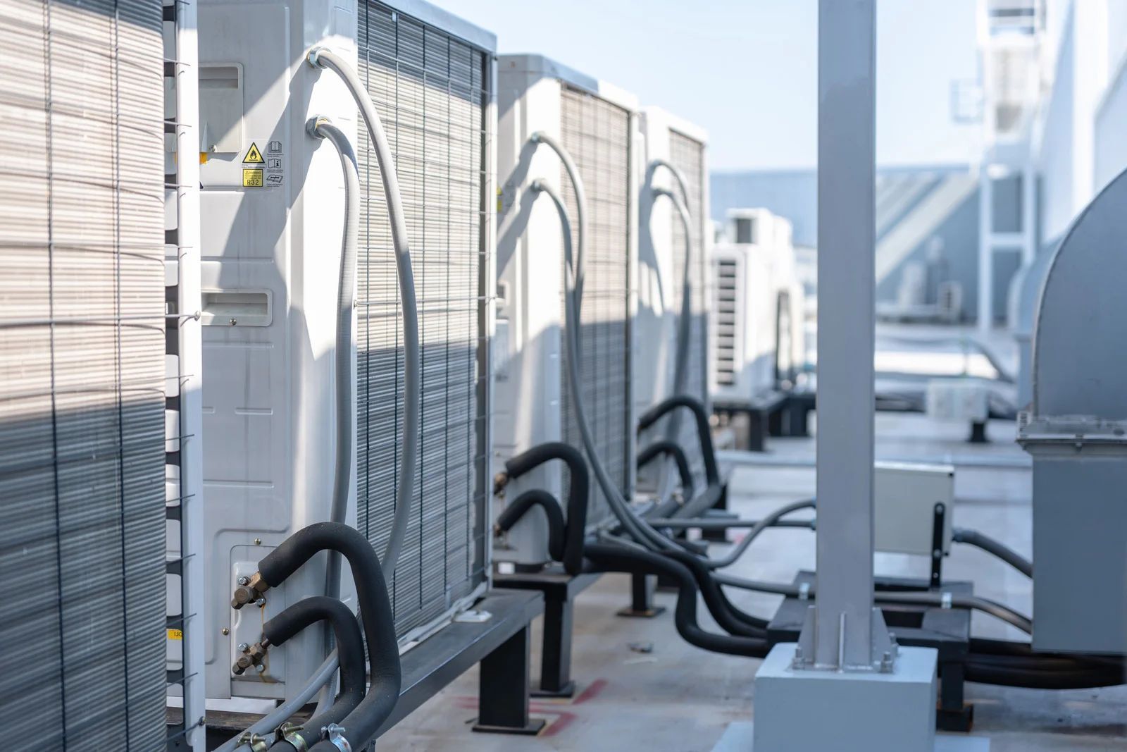 Row of rooftop HVAC units and curved pipes beside a metal fence on a city building rooftop