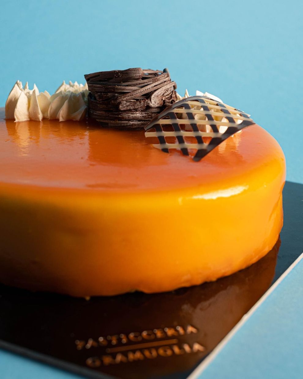 Torta da dessert arancione lucida con decorazioni al cioccolato su base nera e sfondo blu.