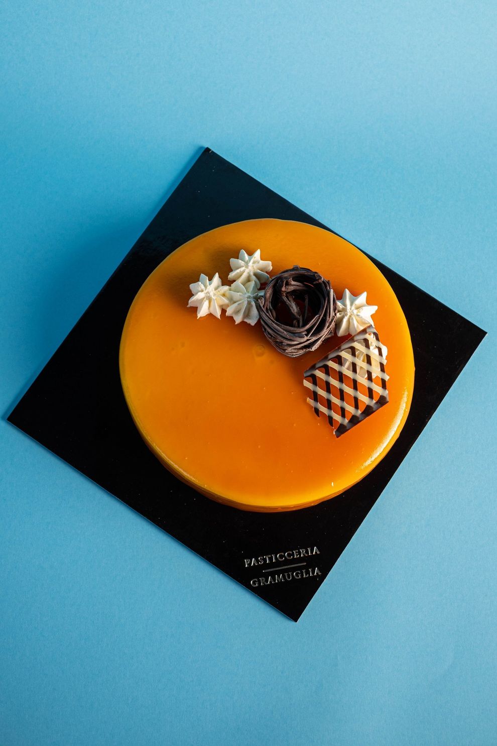 Torta con glassa all'arancia su una base quadrata nera, decorata con cioccolato, fiori e un motivo a scacchiera.