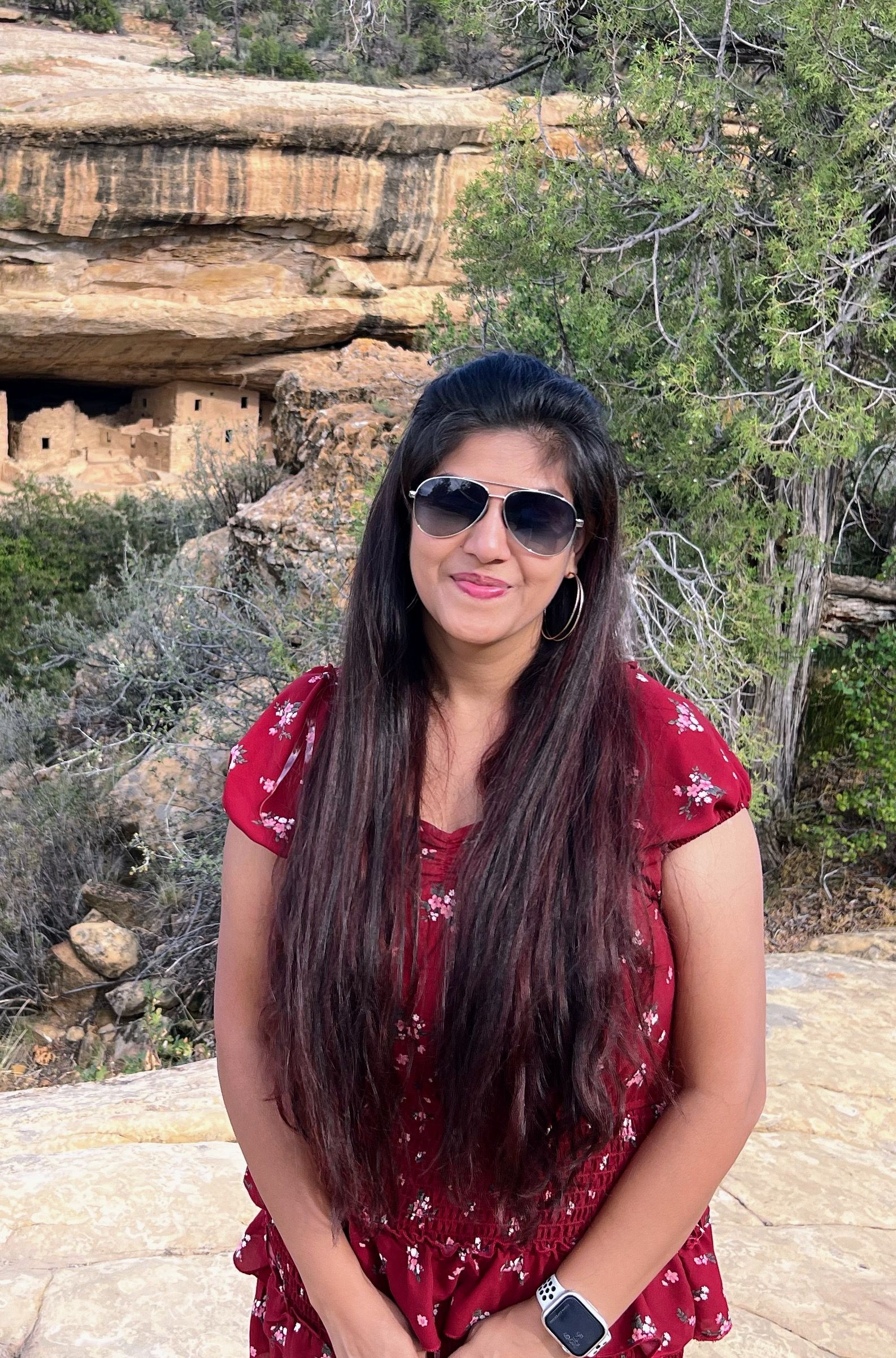 A woman wearing sunglasses and a red top is standing in front of a rock formation.