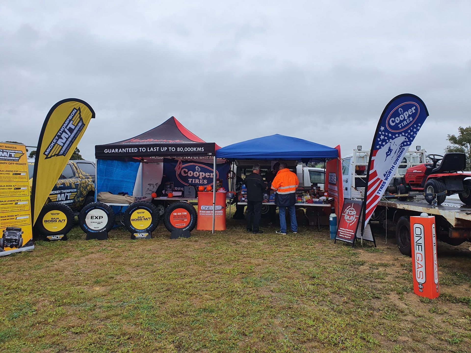 Outdoor event booth with blue and red tents, banners, and people in orange jackets on a grassy field.