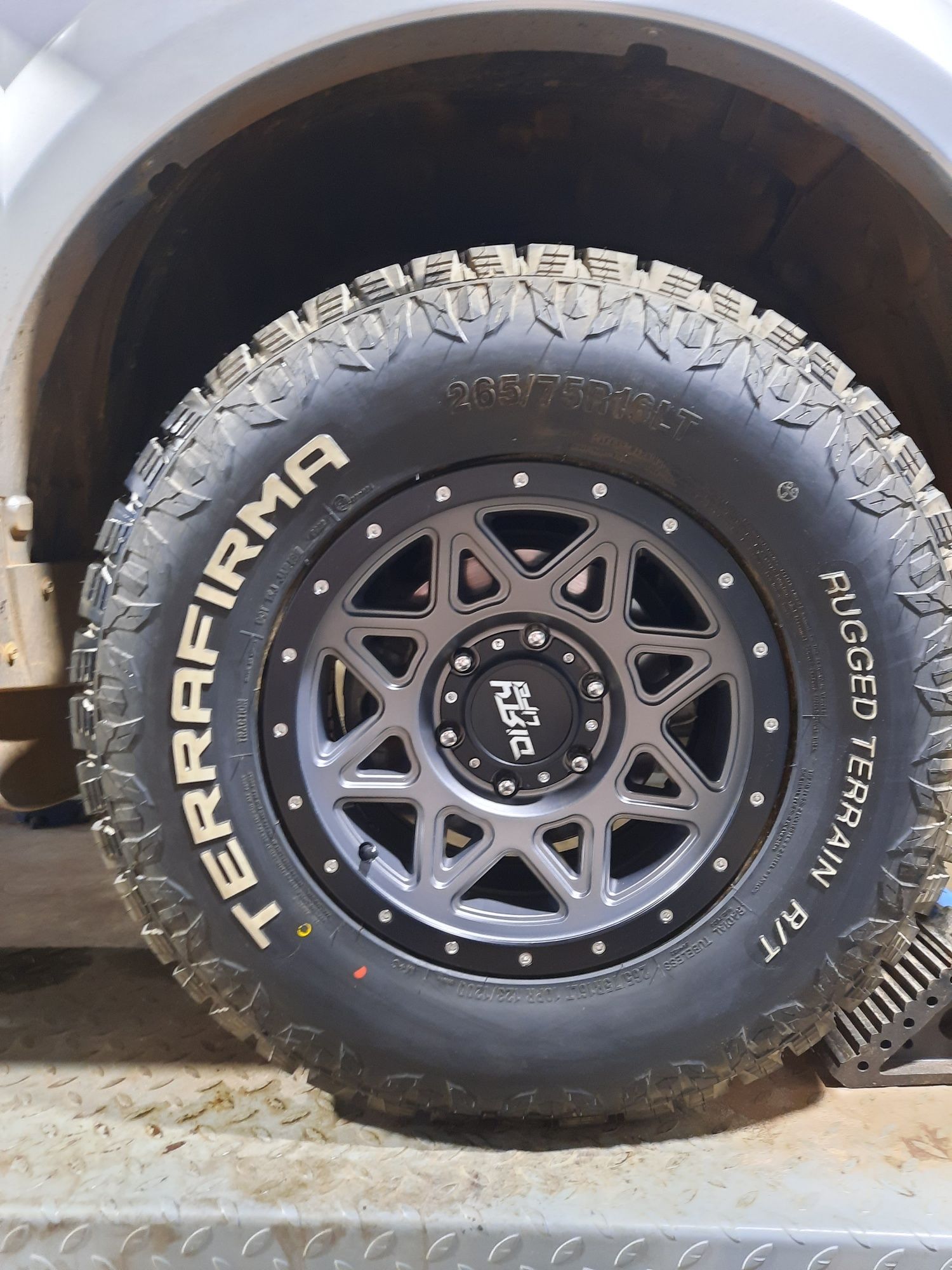 Close-up of a black off-road tire and wheel on a vehicle, with white lettering and muddy tread.