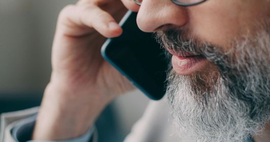 Man with glasses and gray beard holding a smartphone to his ear in close-up.