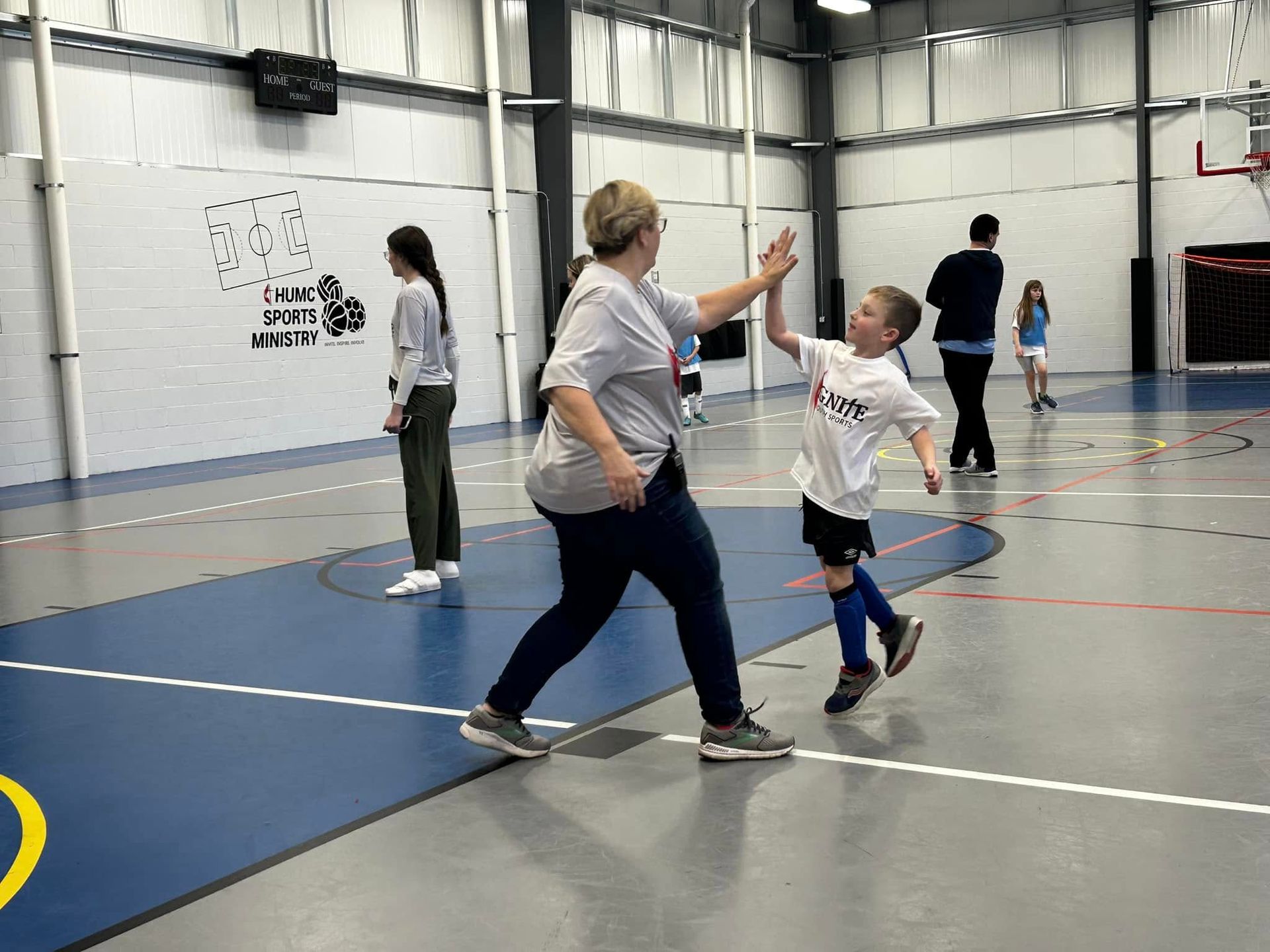 Kids playing indoors in a gym, with one child high-fiving an adult under bright lights.