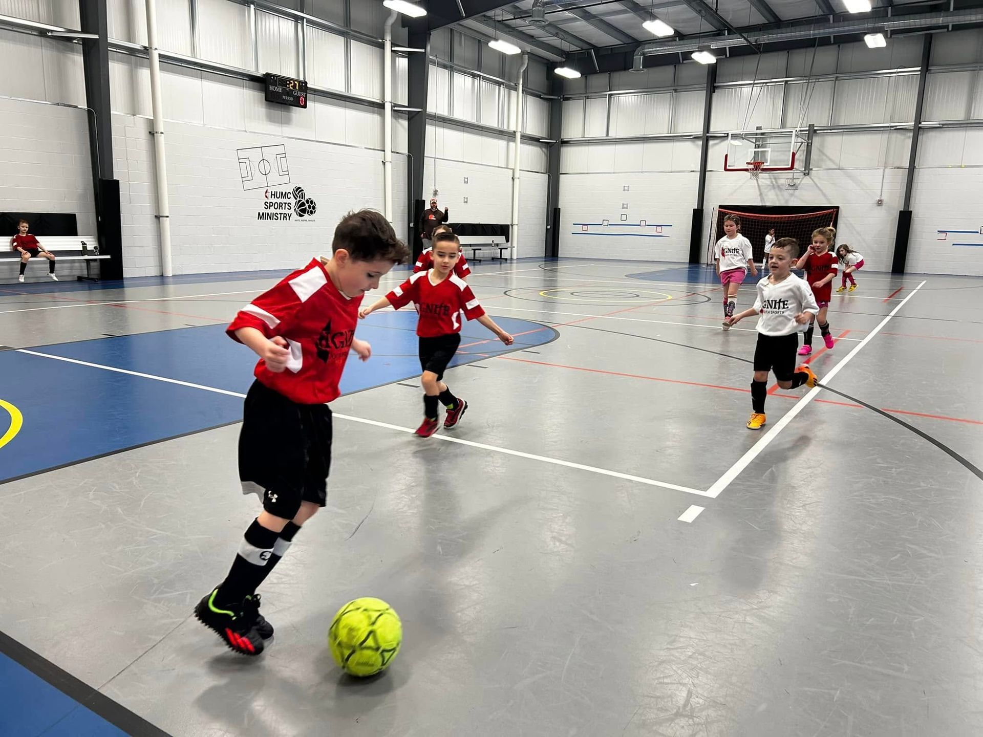 Children playing indoor soccer in a gym, with a boy dribbling a bright yellow ball.