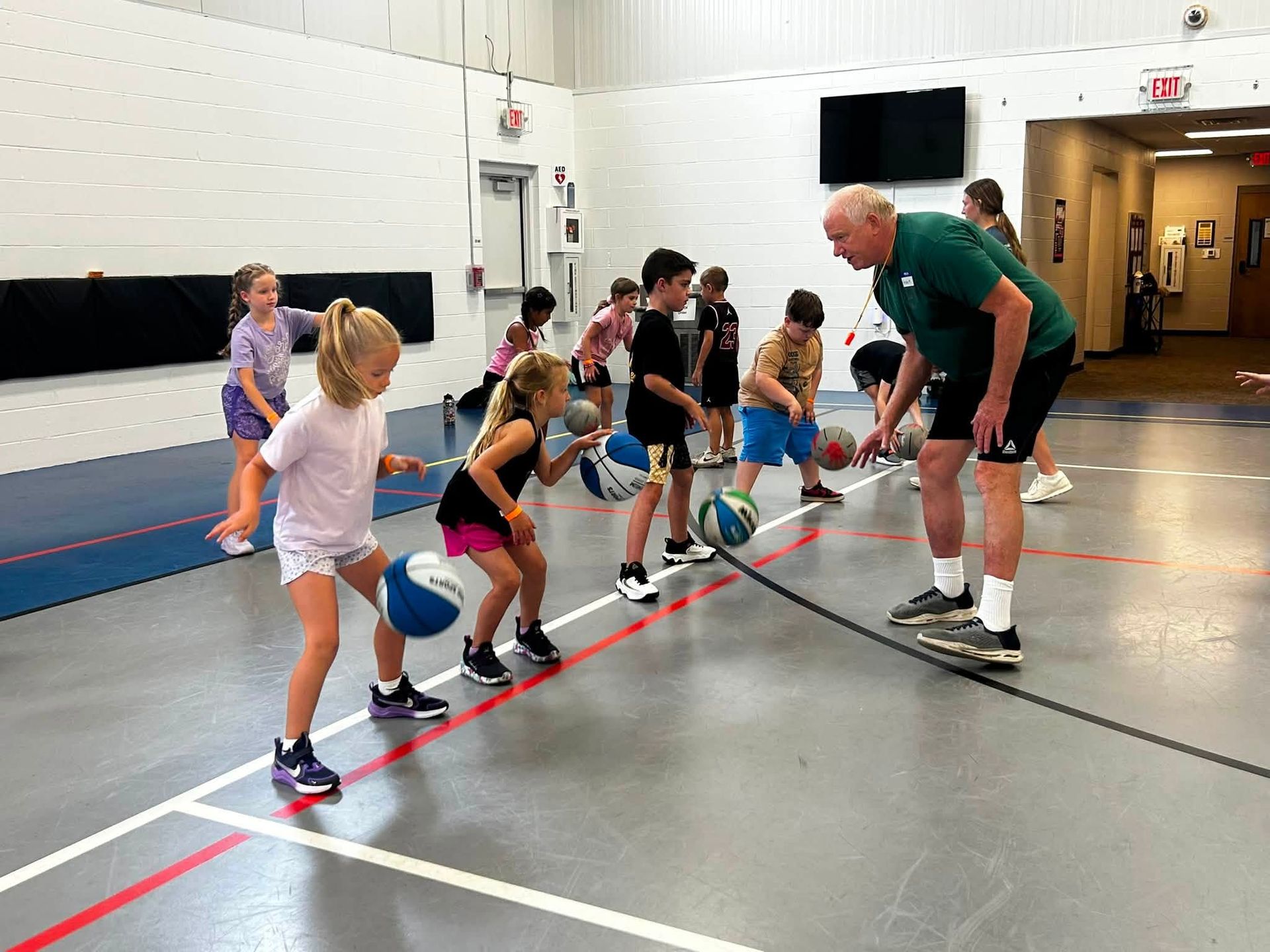 Children dribbling basketballs in a gym while a coach demonstrates stance nearby