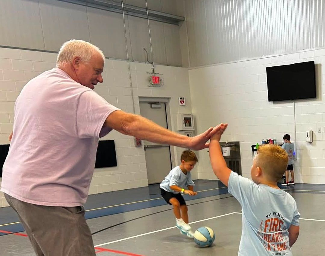 Adult high-fiving a child in a gym while another child dribbles a soccer ball nearby.