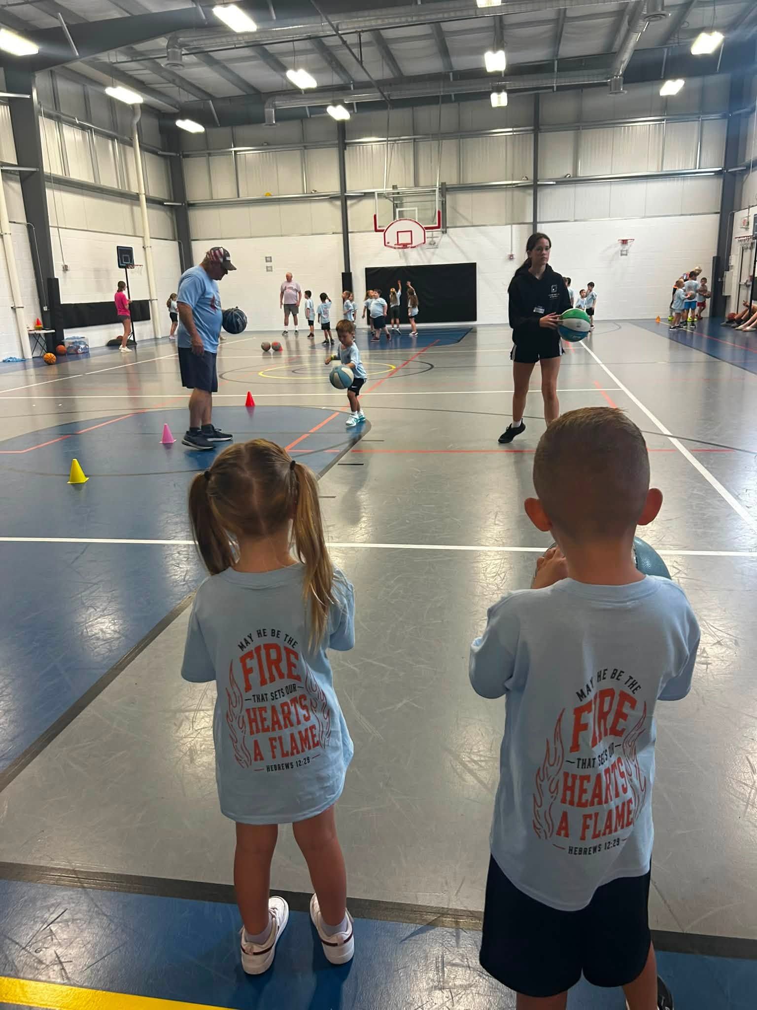 Children watching an indoor gym activity, with cones and people playing in the background.