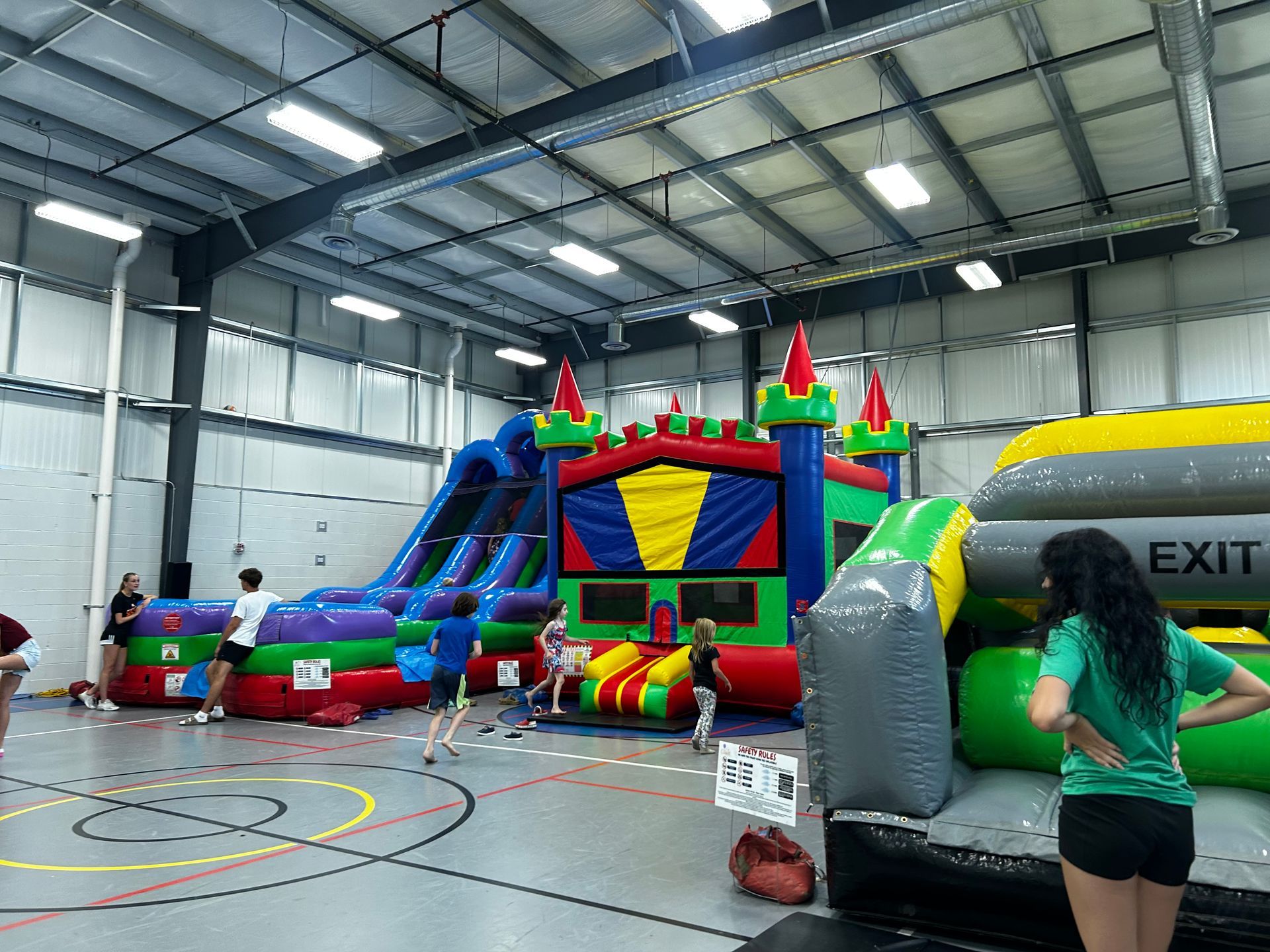 Indoor inflatable obstacle course in a gym, with colorful slides and a green EXIT arch.