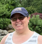 Smiling person in a blue cap and glasses standing outdoors with greenery and rocks behind.