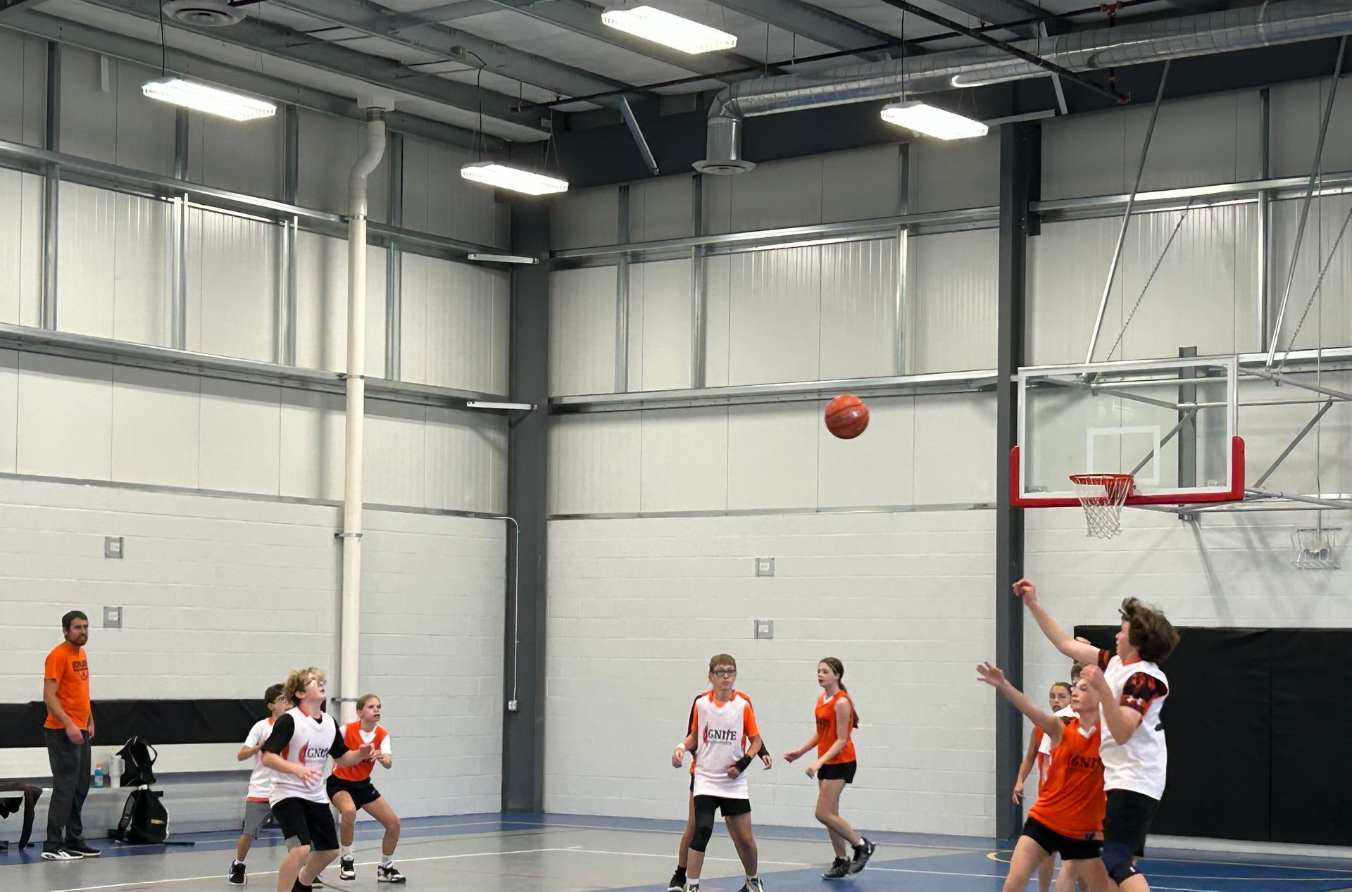Youth basketball game in a gym, players under the hoop as the ball arcs toward the basket.