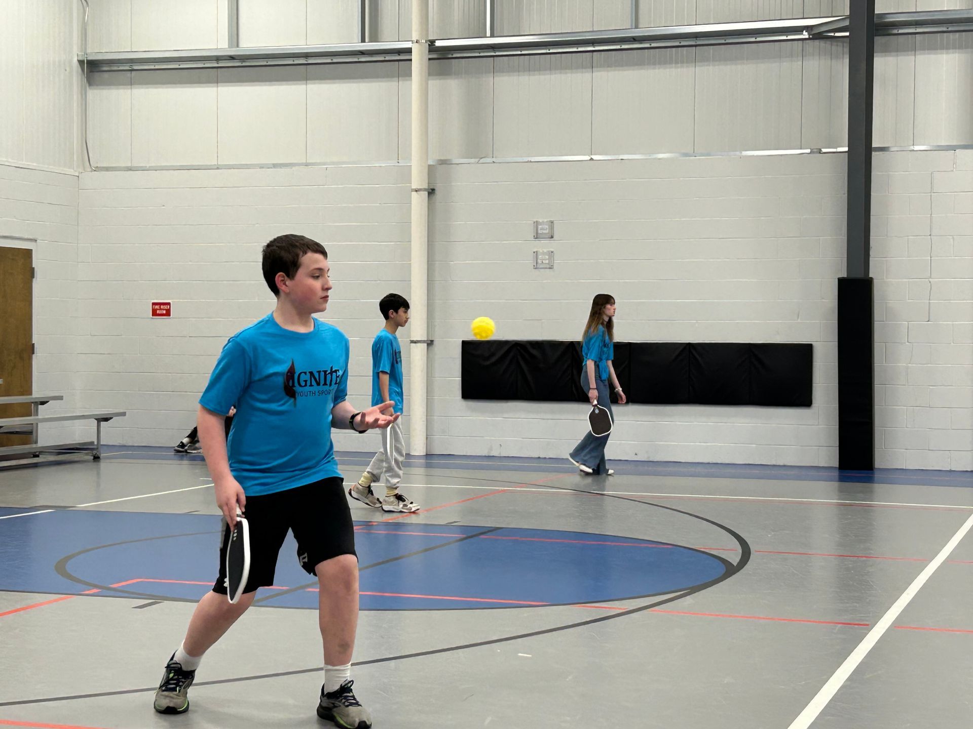 People playing pickleball in a gym, with one player in a blue shirt in the foreground.
