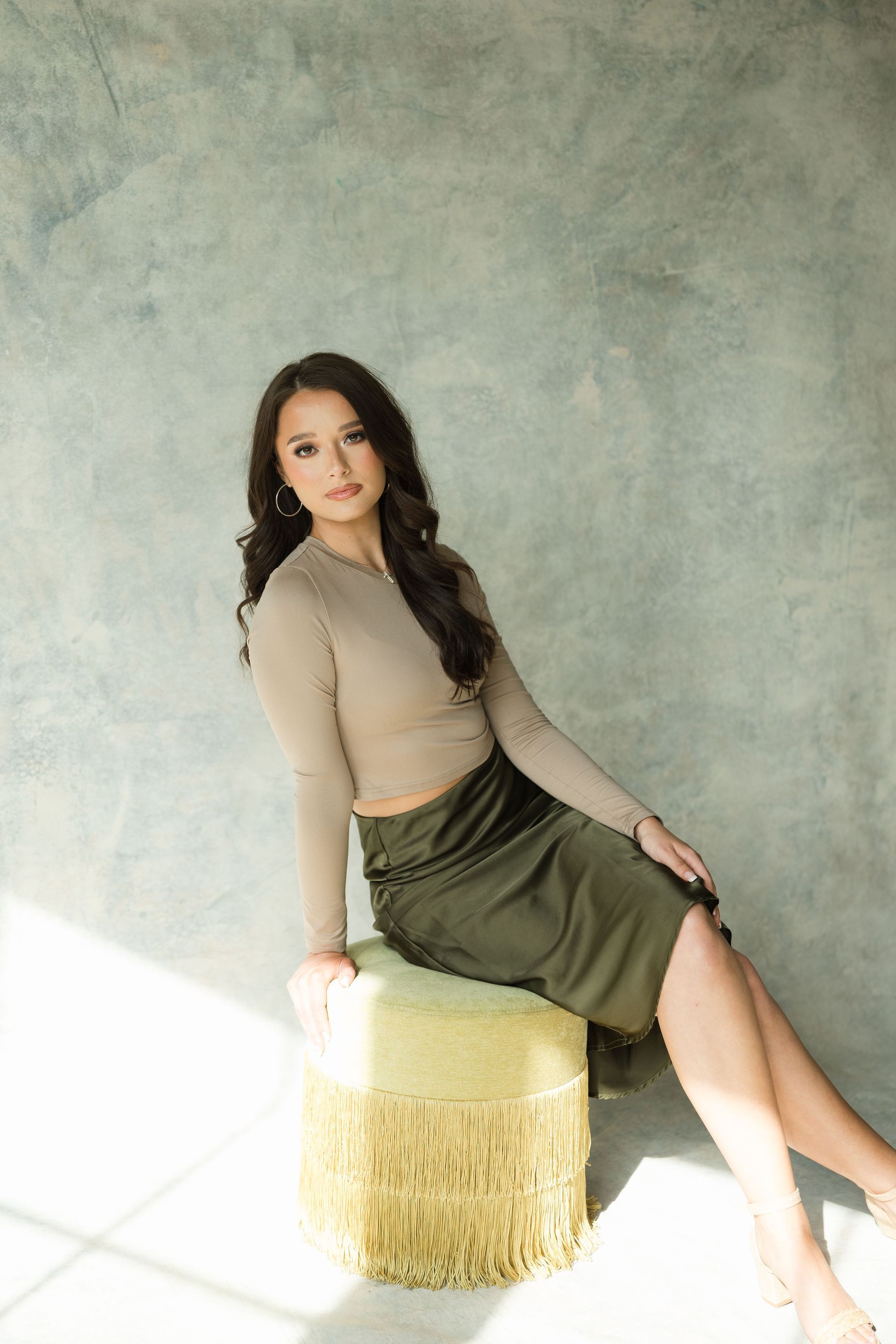 Emily seated on a fringed ottoman against a gray backdrop, wearing a beige top and olive skirt.