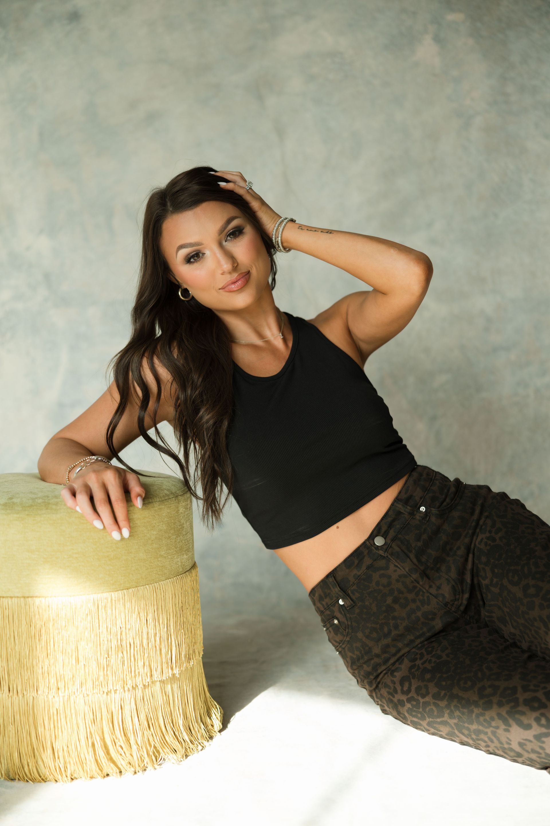 Britney reclining on a gold chair beside a textured wall, wearing a black top and dark jeans.