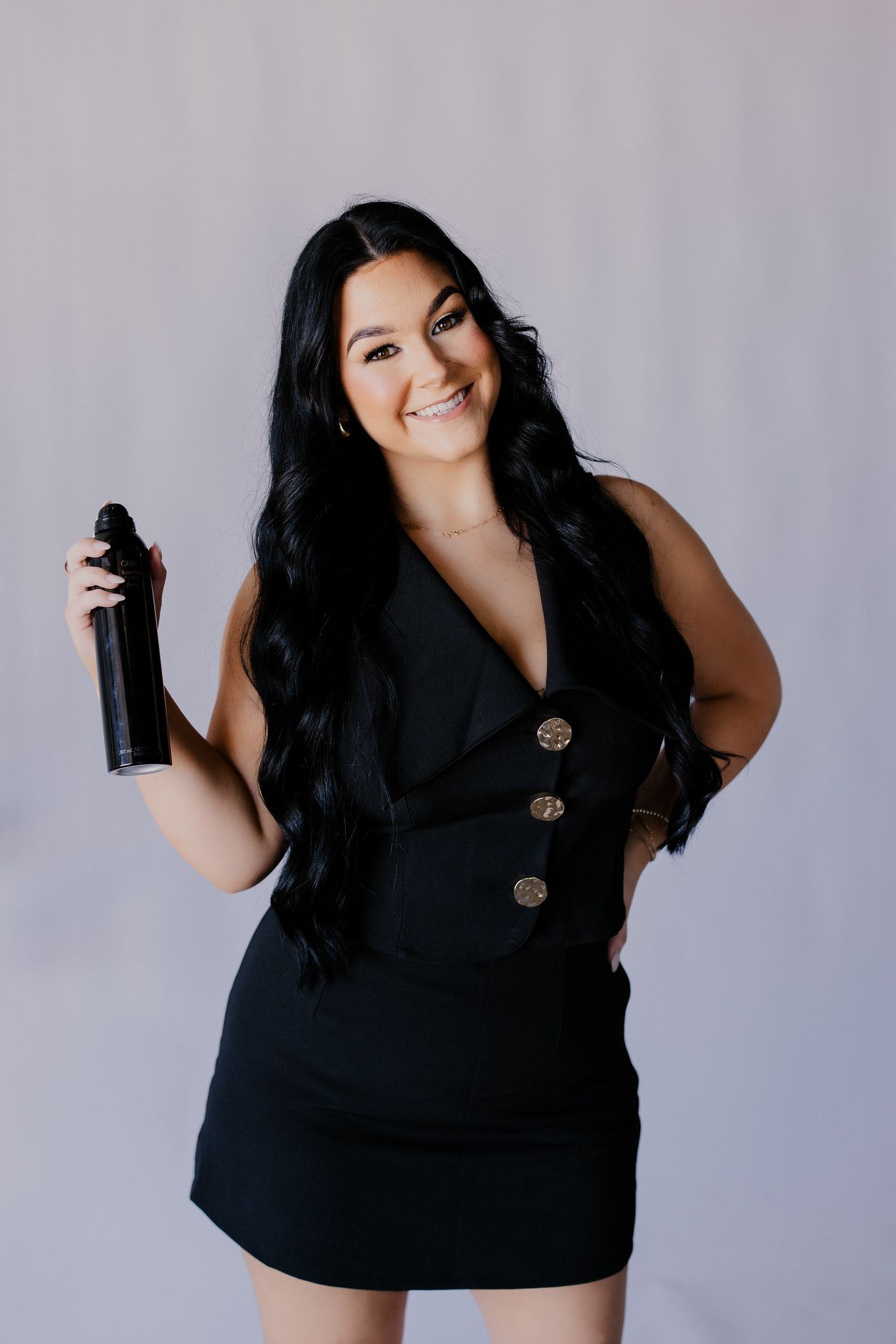 Raven in a black dress smiling while holding a spray bottle against a light gray backdrop