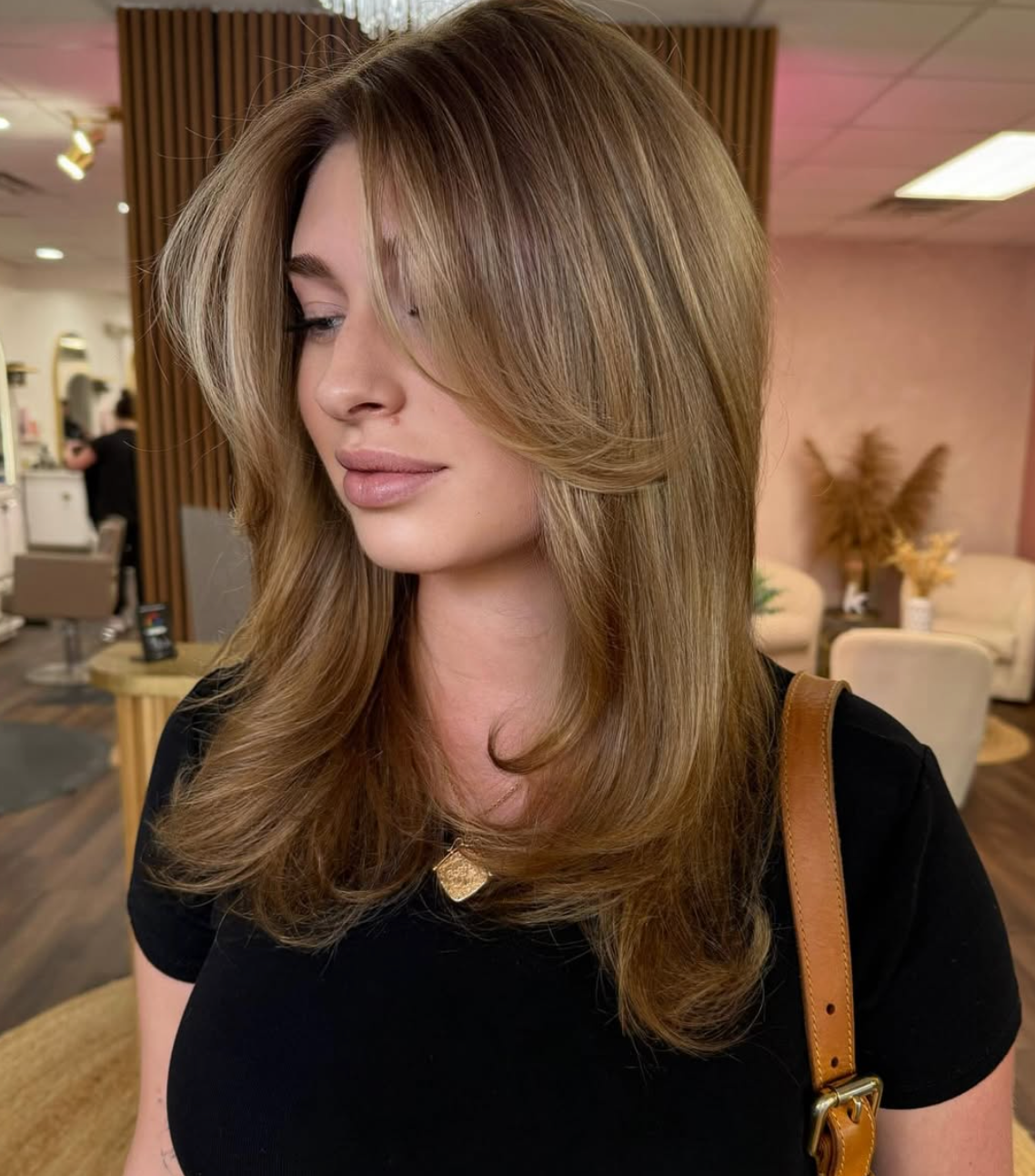 Woman with layered blonde hair in a salon, wearing a black top and tan shoulder bag.