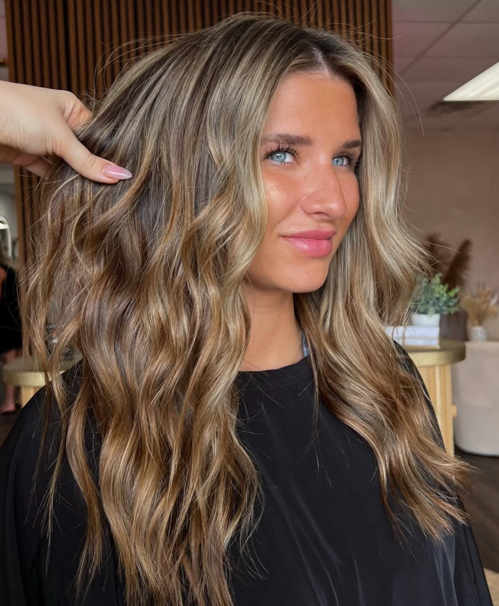 Woman with long wavy blonde hair in a black top, smiling in a salon-like setting.