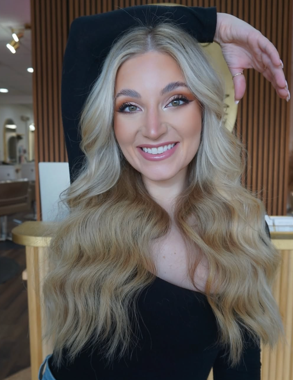 Smiling woman with long blonde hair in a black top, posing indoors with one hand raised behind her head