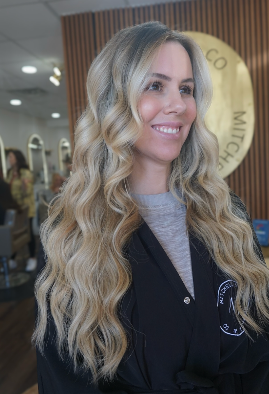 Smiling woman with long blonde waves in a black cardigan indoors, with a mirror and wood-paneled wall behind her