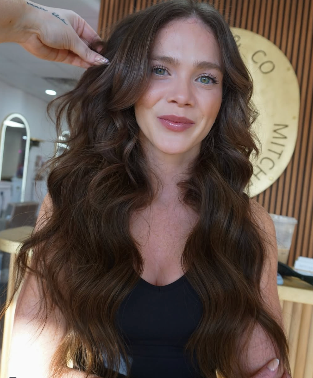 Woman with long wavy brown hair in a black top, smiling indoors with a gold circular sign behind her.